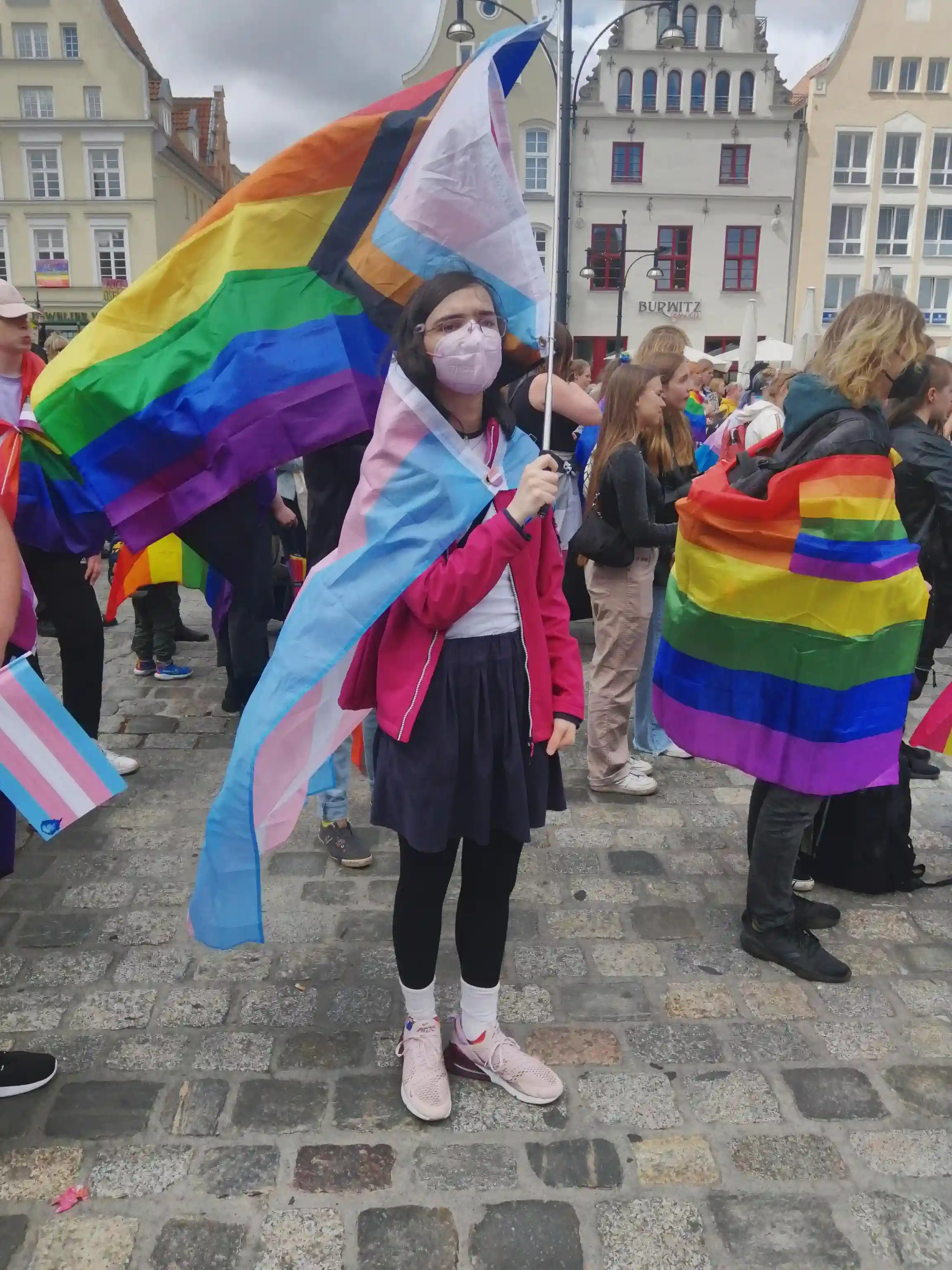 Us, wearing pink jacket, a pink FFP2 mask, and a transgender flag as a cape, held together by a lesbian flag pin, holding a waving progress flag (with a polyamorous flag on the other side, not visible in the picture)